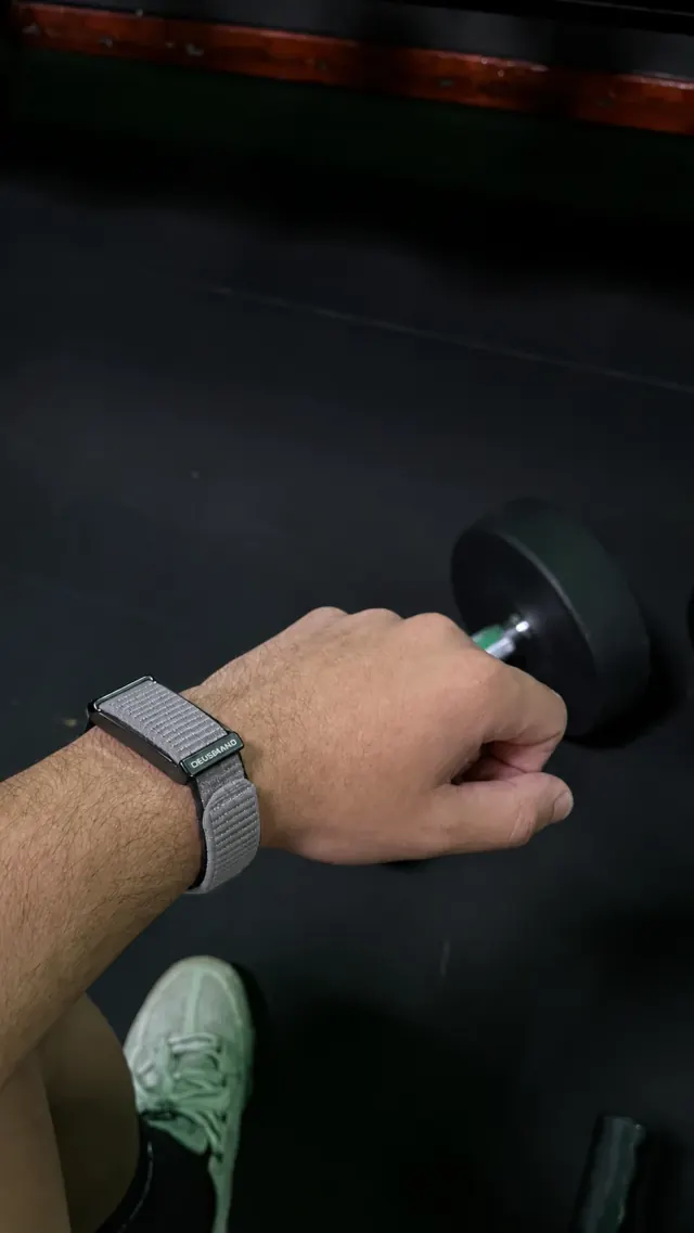 DEUS Band con correa de nylon gris en la muñeca, entrenando con mancuernas en el gimnasio. Foto del cliente editada para mostrar la correa gris